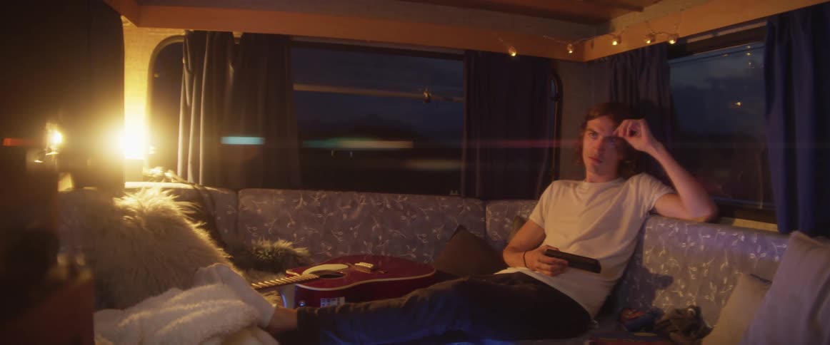 Man with long hair and white shirt watches television on board at dusk