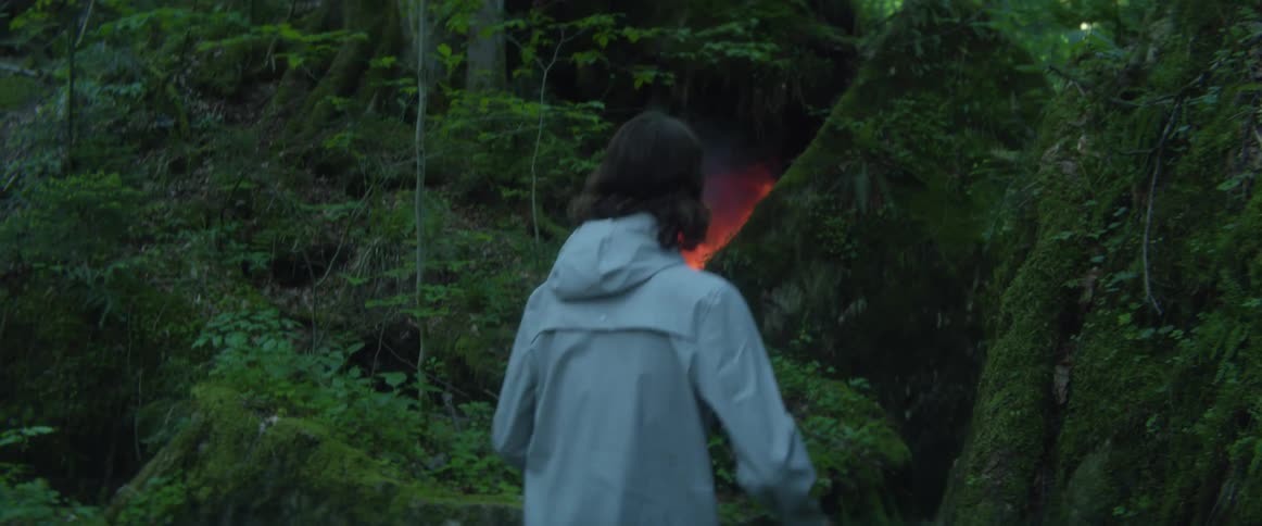 A teenage boy in a forest walks towards what appears to be a fire in a cave