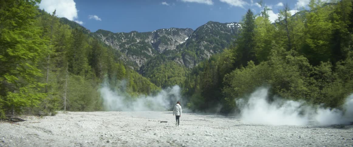 Man with gray coat and black pants walks along valley between mountain forests