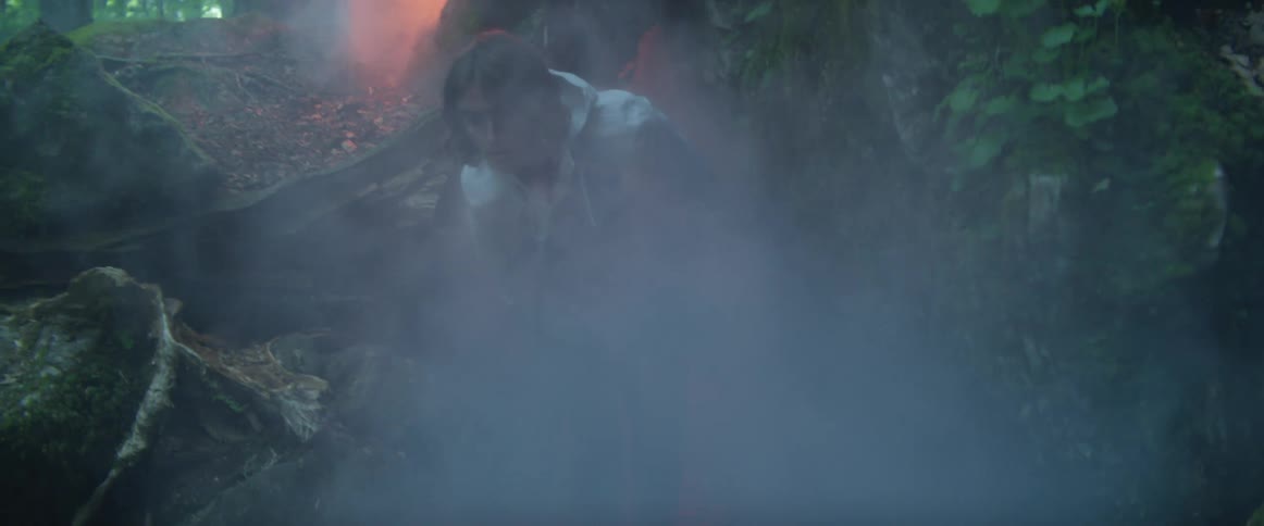 Man with long hair and gray raincoat comes out of the smokey cave in the woods
