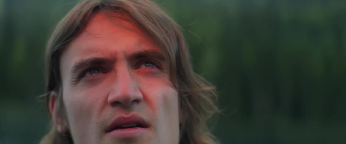 A man looks up as an orange light shines on his face as he stands near a forest