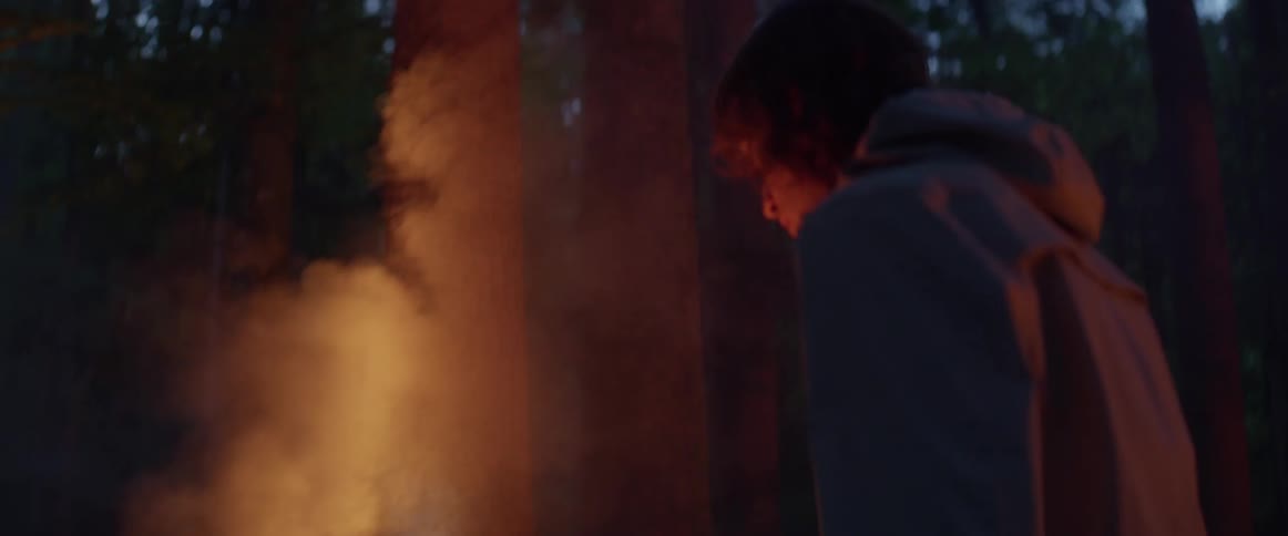 Man with long hair and jacket reaches his hand out to smoky campfire light in the dark forest