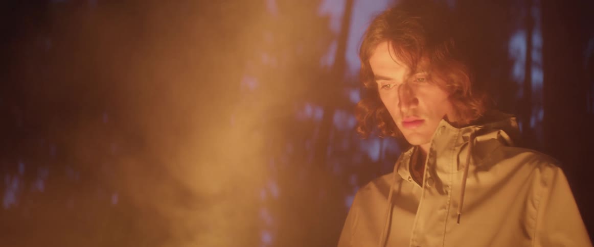 A teenage boy in a forest stands walks towards a fire and looks in to it