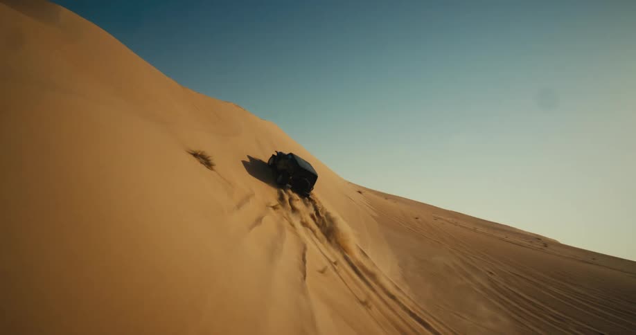 A black truck travels up a desert hill as sand flies behind it