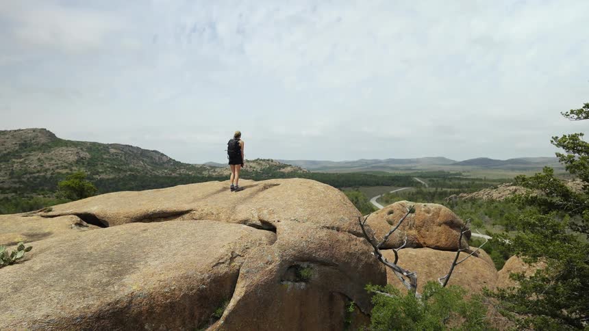 A female hiker looks out over the countryside while standing atop a rock formation in the wilderness
