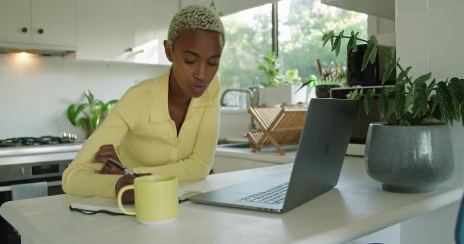 A woman is talking on a video call on a laptop and writing things in a notebook