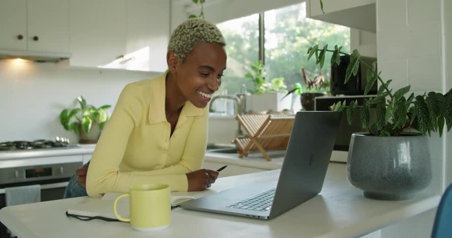 A woman is having a video chat using her laptop while standing in her home kitchen during the daytime