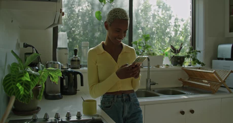 A woman stands in her kitchen and smiles and laughs as she scrolls on her cell phone