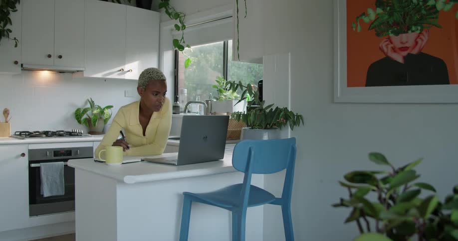 A woman in a kitchen with a pen in front of a laptop with a wall and stove in the background