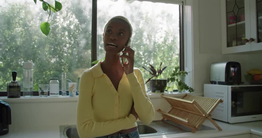 A woman in a room talking on a smart phone with a wall and window in the background