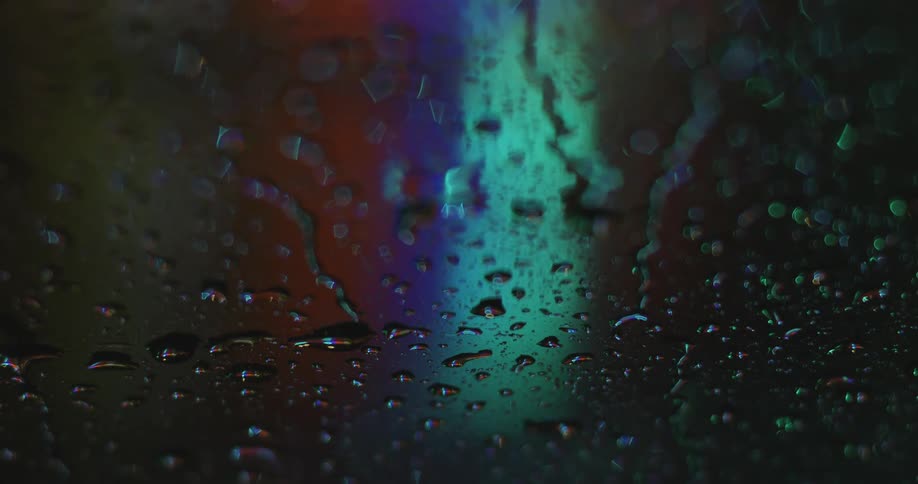 Water droplets slide down glass at night with colored lights behind