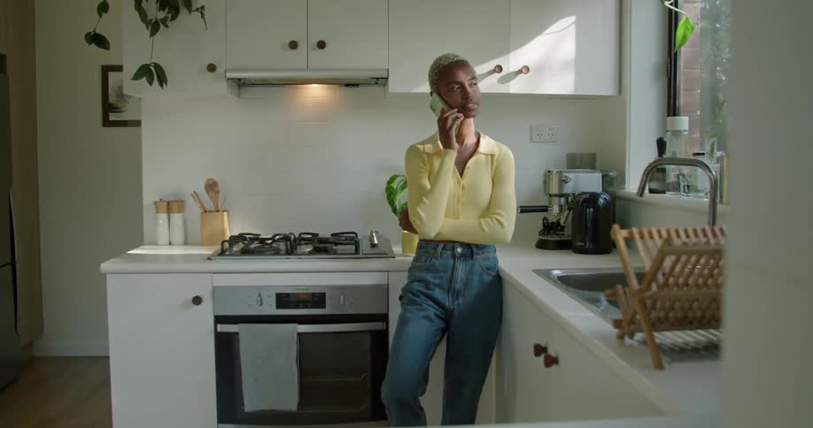 A woman is standing in a kitchen smiling as she talks on her cell phone