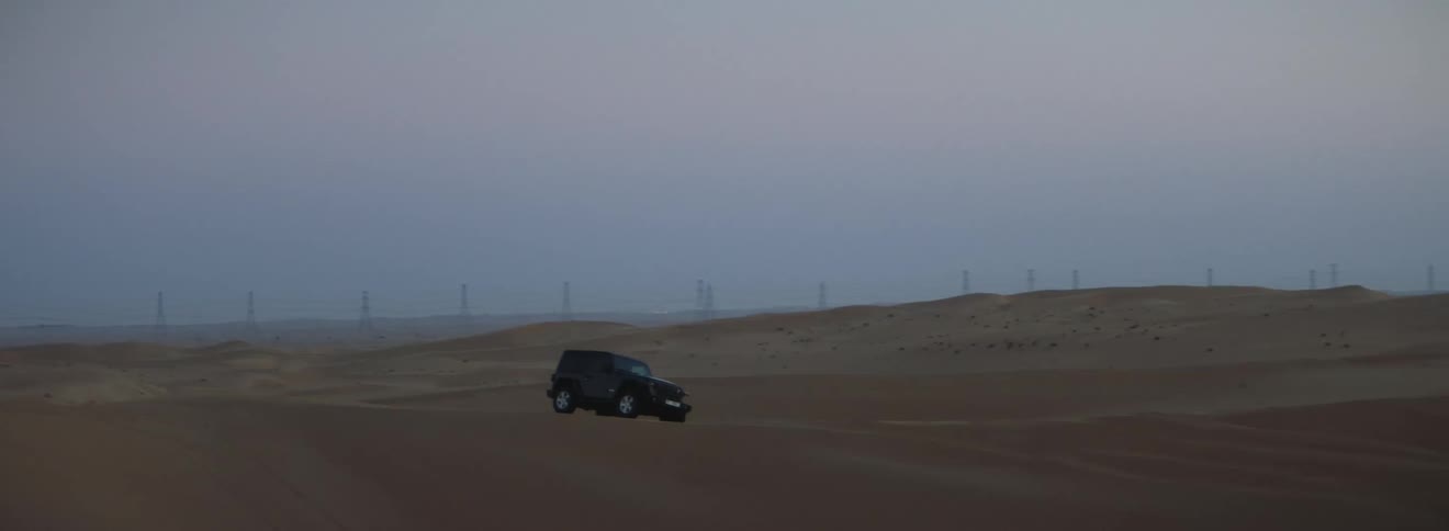 A black jeep travels across a desert and utility towers are in the distance