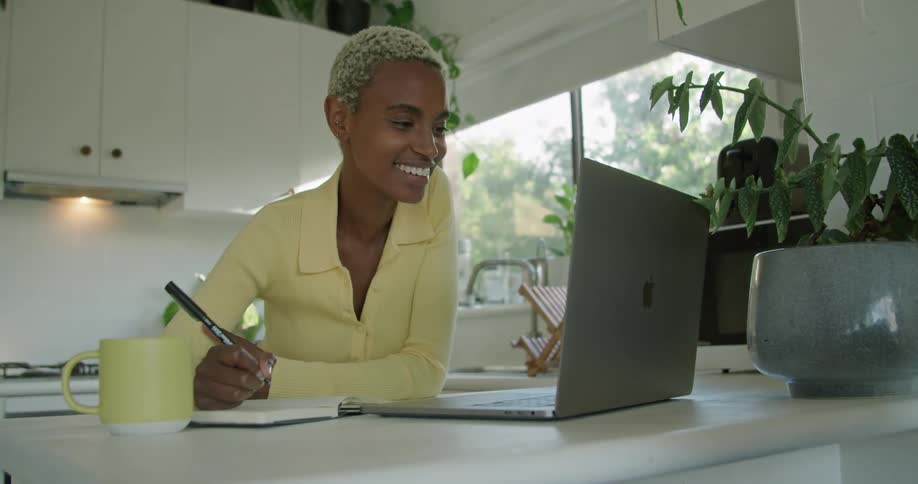 A woman is holding a pen and writing in a book as she is having a video conversation with someone on a laptop computer