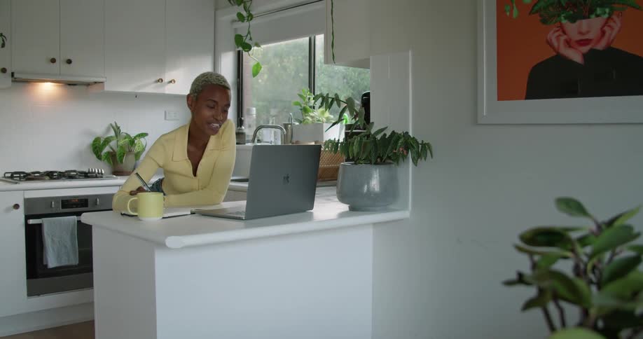 Woman takes notes working at kitchen counter and participating in video conference