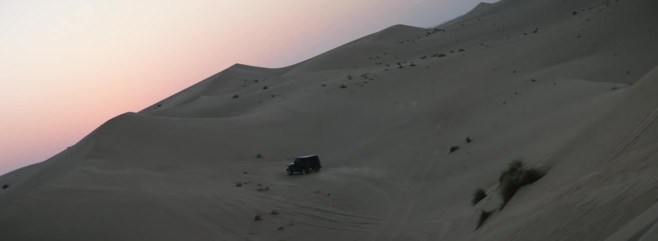 A vehicle travels off road across a tall sand dune in the evening