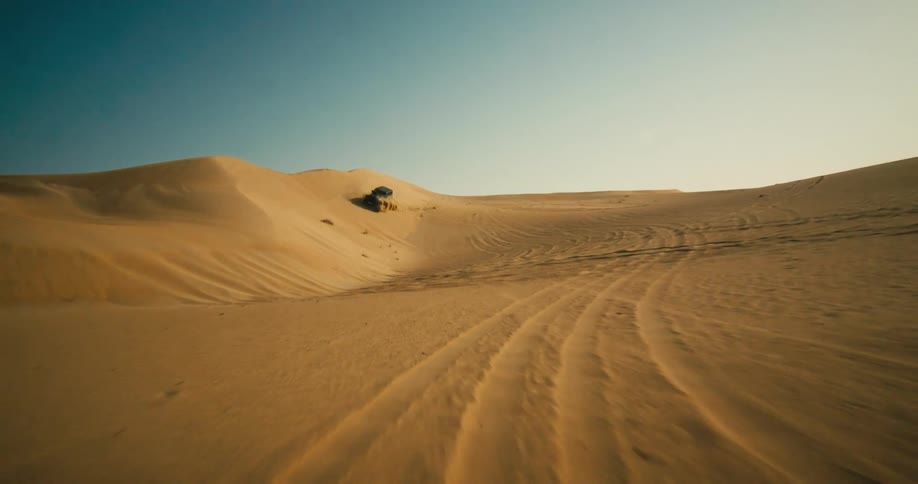 Jeep drives sliding along side of sand dune kicking up sand under clear blue sky near track covered desert surface