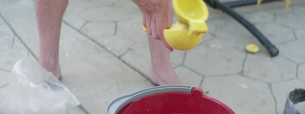 A man is squeezing lemon juice from lemons into a bucket while outdoors on a sidewalk during the daytime
