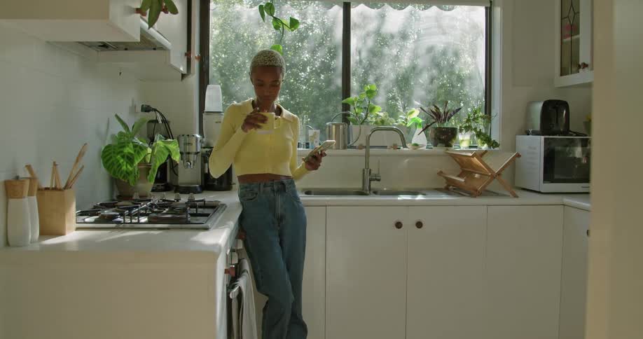 A woman is standing in her kitchen drinking coffee and texting on a smartphone in the daytime