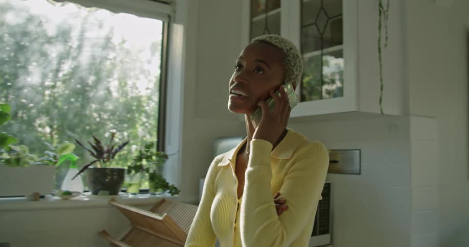 A woman is smiling and talking on a cell phone in her kitchen by a sunny window
