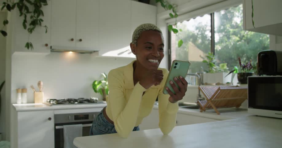 Woman with short hair yellow vest and jeans leans on kitchen island and makes video call on smartphone in the kitchen