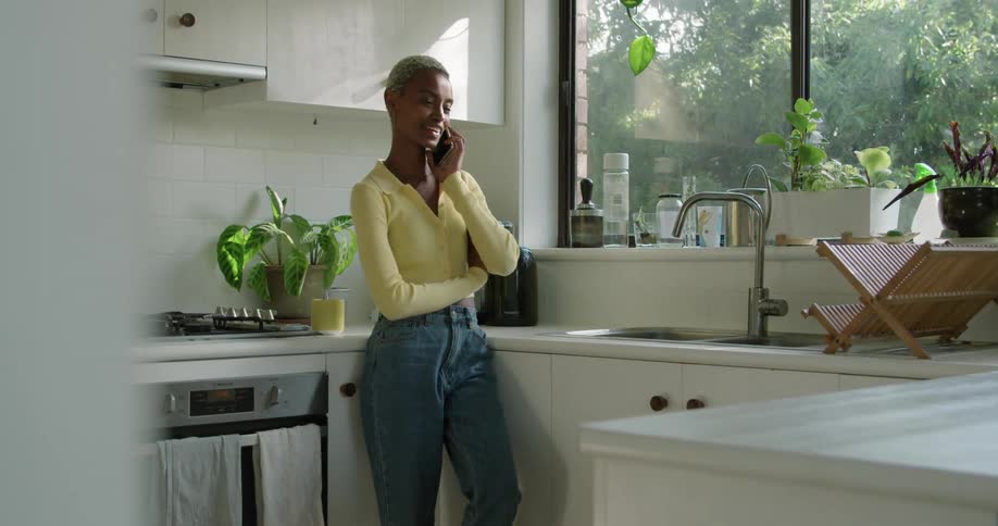 A woman in a kitchen talking on a smart phone with a wall and window in the background