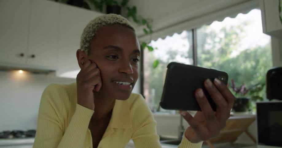 A woman is sitting in the kitchen using her phone to video chat with someone