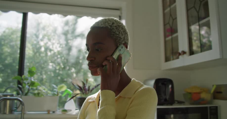 A woman in a room talking on a smart phone with a wall and window in the background