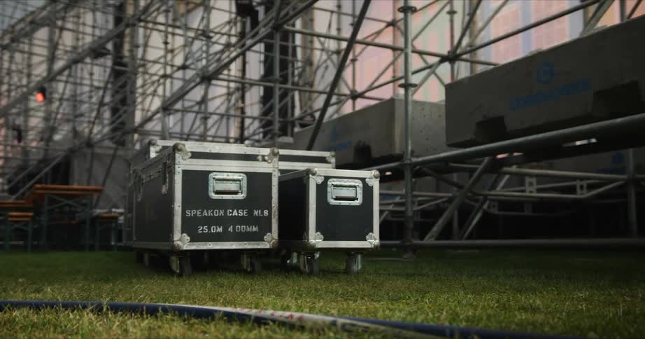A man in jeans and boots walks past equipment trunks and a power cable near an event stage set up