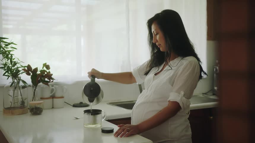 A pregnant woman in a kitchen pouring hot water from a kettle into a coffeemaker