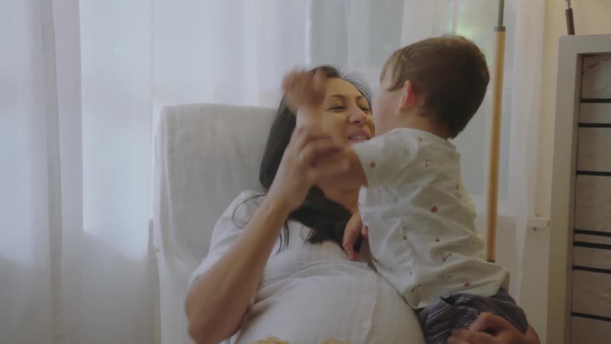 Pregnant mother with black straight hair and white shirt sits in armchair playing with her son