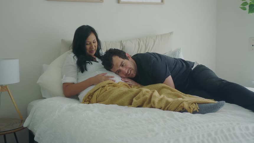 A pregnant couple are relaxing in a bed at home while the man listens to the womans stomach during the day