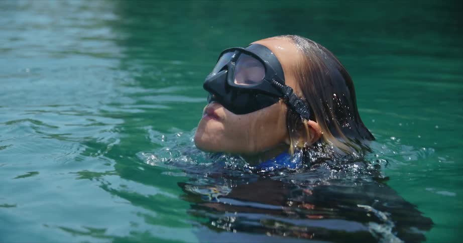A woman wearing swimming goggles is underwater and she lifts her head up from underwater