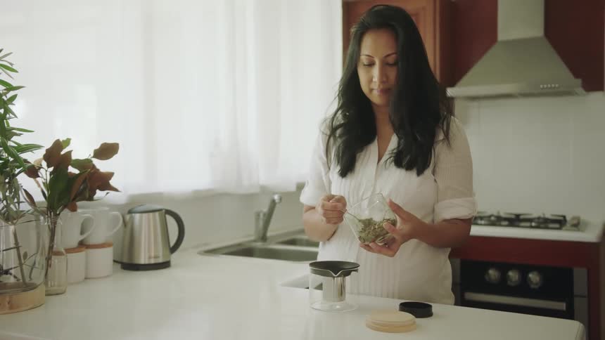 A pregnant woman is in the kitchen scooping tea leaves into a steeper jar