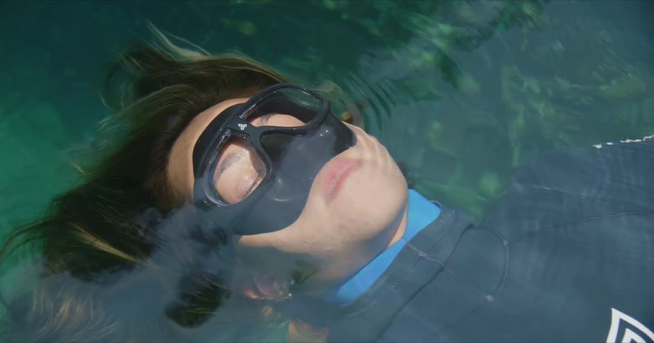 Woman with brown hair black mask and wetsuit lies on back in water