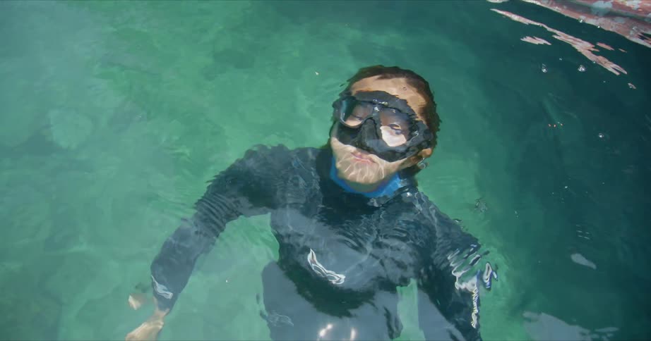 A woman in a diving suit and face mask is emerging from underwater in a pool during the daytime