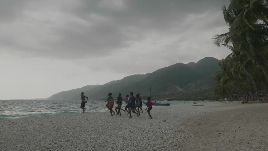 A group of children are running toward the ocean water on a beach with a mountain in the background and rain clouds in the sky