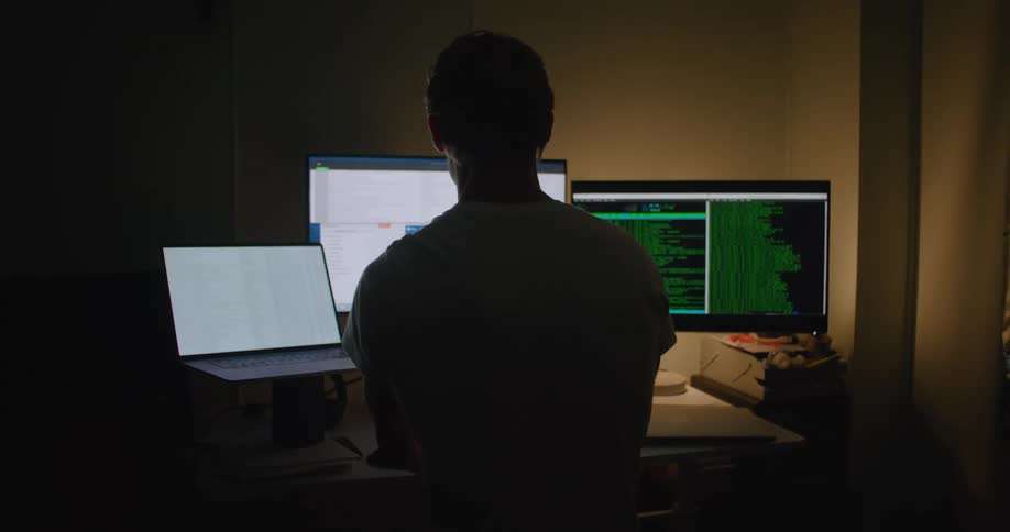 Man with white t shirt sits at desktop and works on laptop and computer in dark room