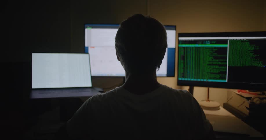 A man is sitting in front of a laptop computer and two other computer monitors
