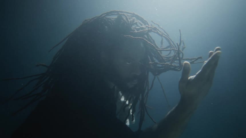 Man with braided hair and beard extends his arms underwater
