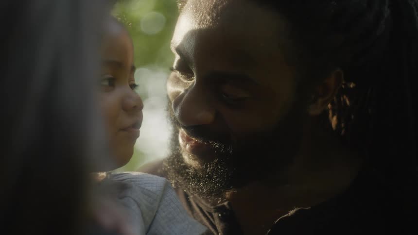 A man kissing a child with a woman in the foreground with trees in the background