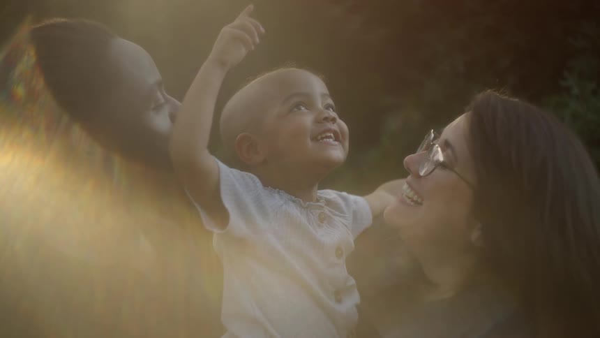 A happy family and their child are outdoors as the child is smiling a pointing upward to the sky in the evening