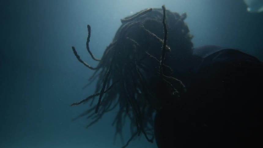 Man with beard and braided hair goes underwater while papers float underwater