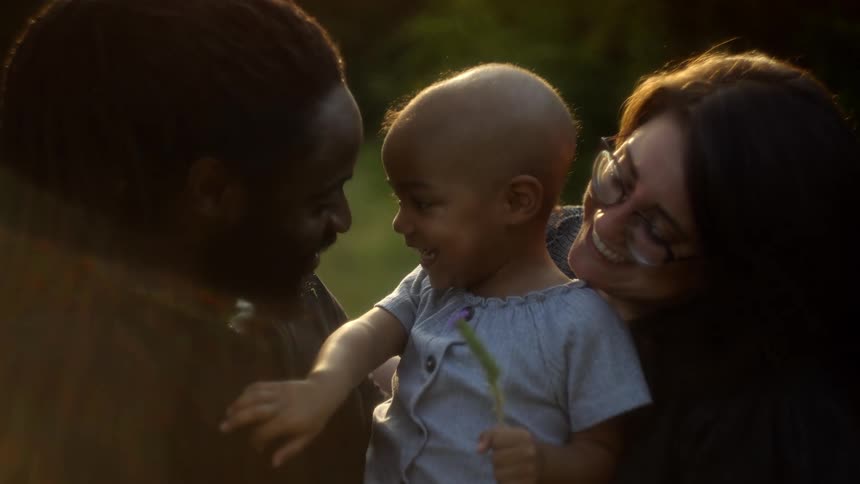 A mother and father are smiling and holding their toddler girl outside