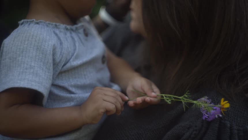 A woman is holding a toddler girl who is holding some wild flowers