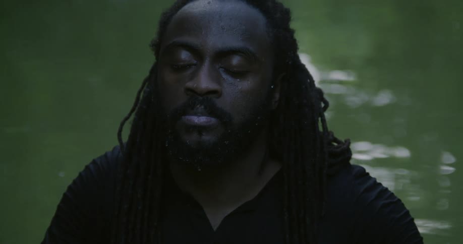 Man with beard braided hair and black shirt sits in stream with eyes closed