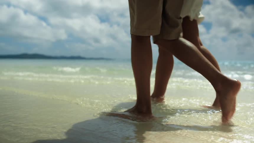 Man and woman with brown shorts and hat walks along the tropical beach on party cloudy day