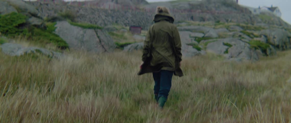 A woman wearing a coat is walking through tall grass toward a lighthouse on a rocky hill