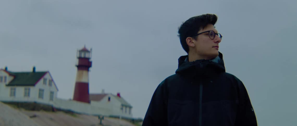 A man with glasses standing outside with a lighthouse the sky and clouds in the background