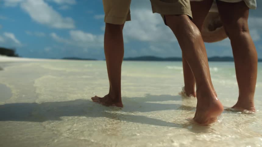 Man and woman with shorts and hat walk along the white sand beach next to the sea